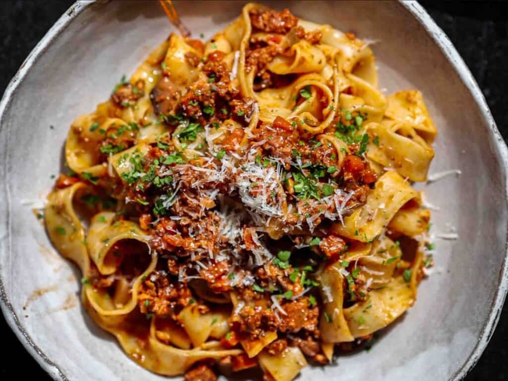 A bowl of pappardelle pasta topped with a rich vegan bolognese sauce, garnished with grated vegan parmesan cheese and chopped parsley.